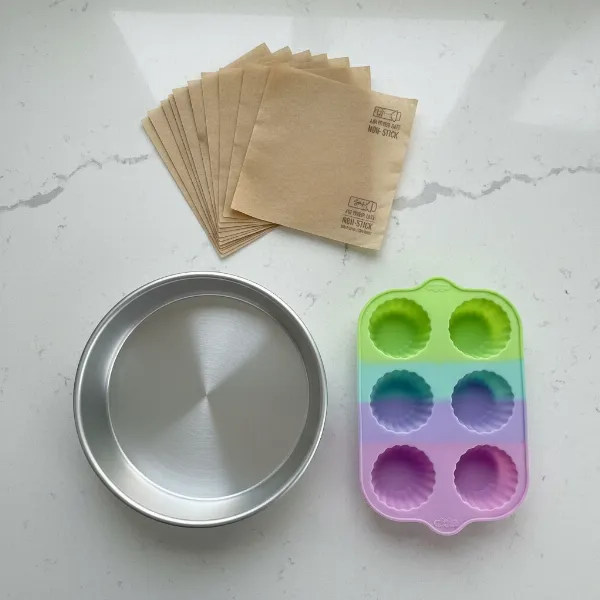 Various air fryer safe cookware options including a small metal baking dish, a silicone muffin mold, and parchment paper liners, ready for use.